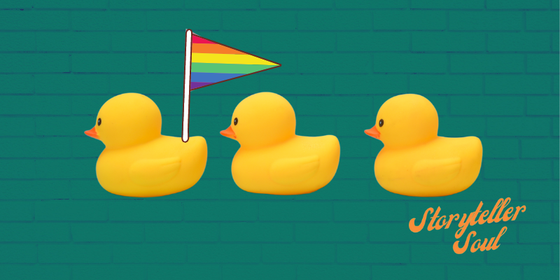 Row of three yellow rubber ducks with the first duck holding a triangular rainbow pride flag.