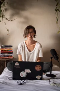 White, middle-aged woman with chin-length brown hair sitting on a bed in front of her laptop computer with a microphone at her side and a stack of books behind her.