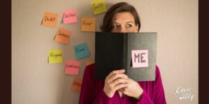 White woman with brown hair holding a black book in front of her face that says "me" with colorful post-it notes filled with emotion words around her.